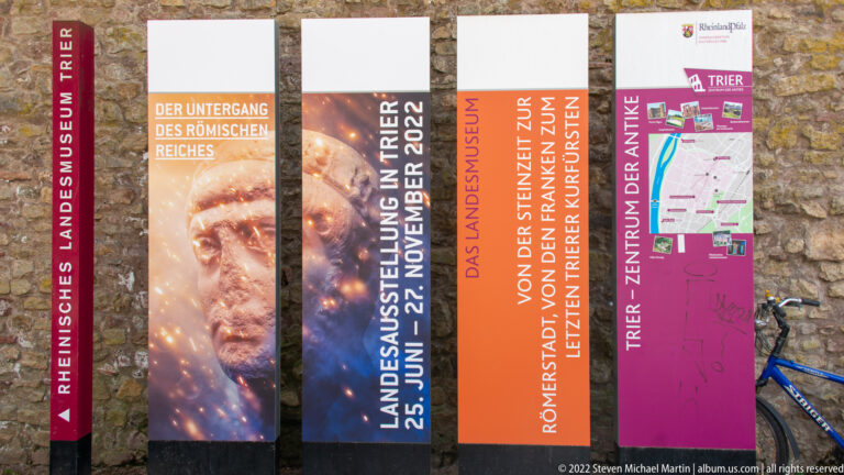 Vertical exhibition banners for the Rheinisches Landesmuseum Trier mounted against a stone wall, including Roman-history exhibition text.
