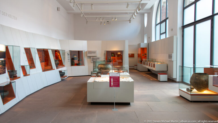 Bright archaeological museum gallery with glass display cases, pottery, angled white panels, and tall windows.