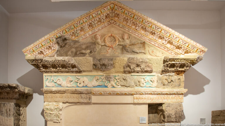 Ancient stone pediment and carved entablature with surviving red, blue, and yellow painted decoration in a museum display.