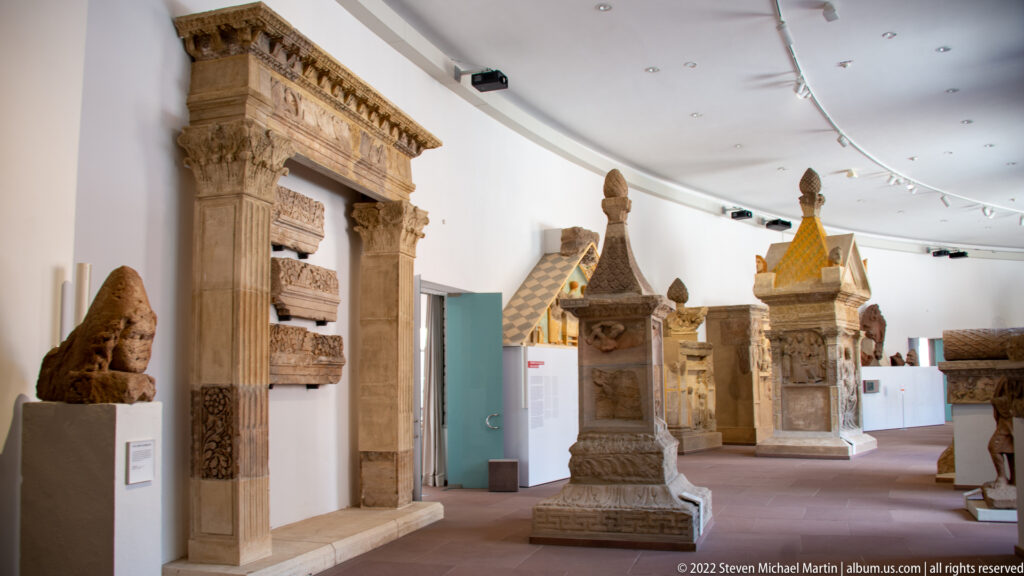Gallery of Roman stone monuments with tall funerary structures, carved reliefs, doorway fragments, and a lion sculpture.