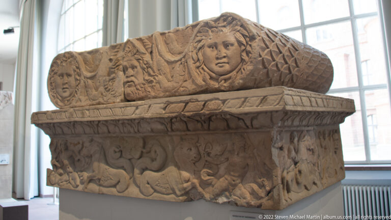 Roman stone sarcophagus with carved figures, garlands, animals, and a gabled lid centered on a large face.
