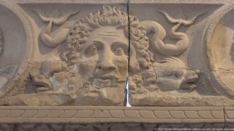 Close detail of a carved Roman face with curling hair, serpentine forms, and weathered stone surface.