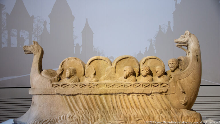 Boat-shaped Roman stone monument with several human figures, circular forms, and animal-headed ends.