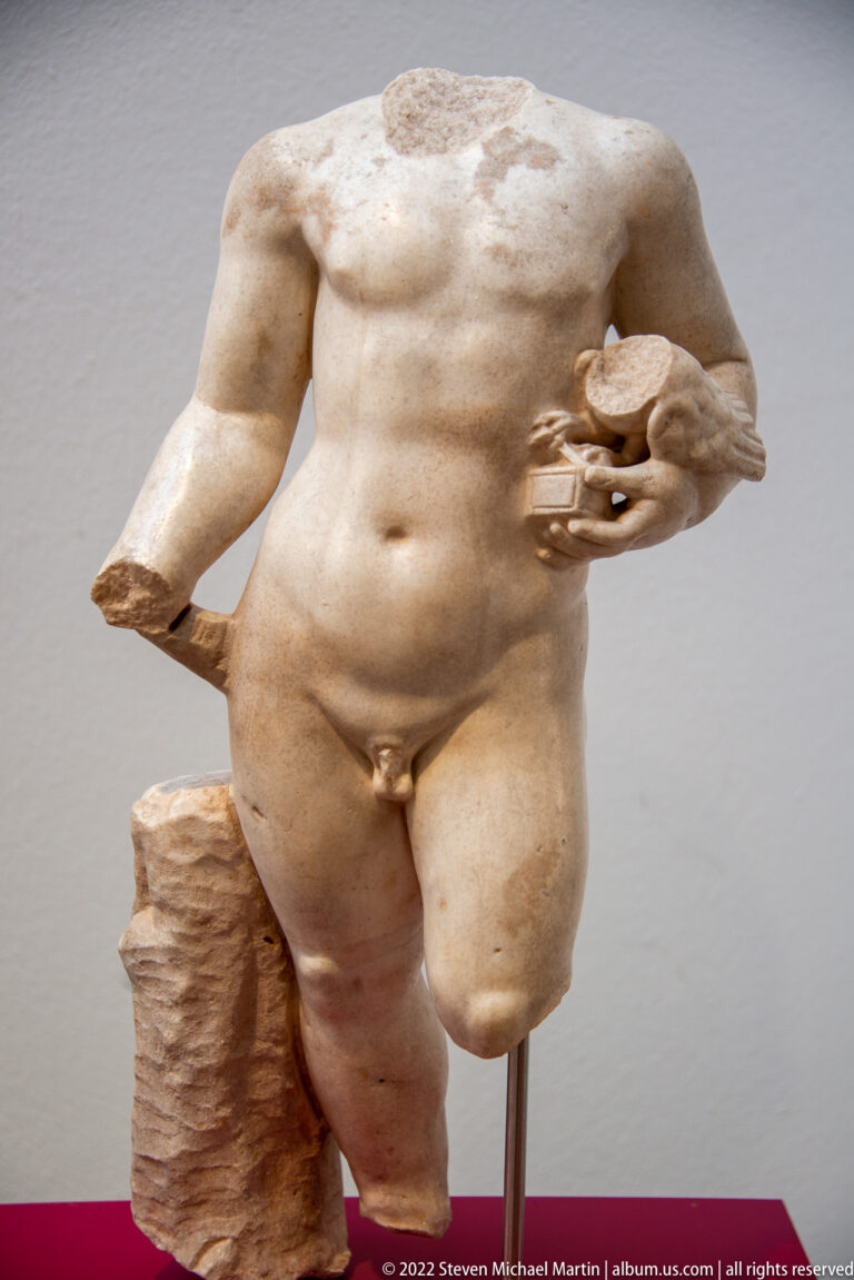 Fragmentary nude male Roman statue with missing head and limbs, one hand holding a rounded object.