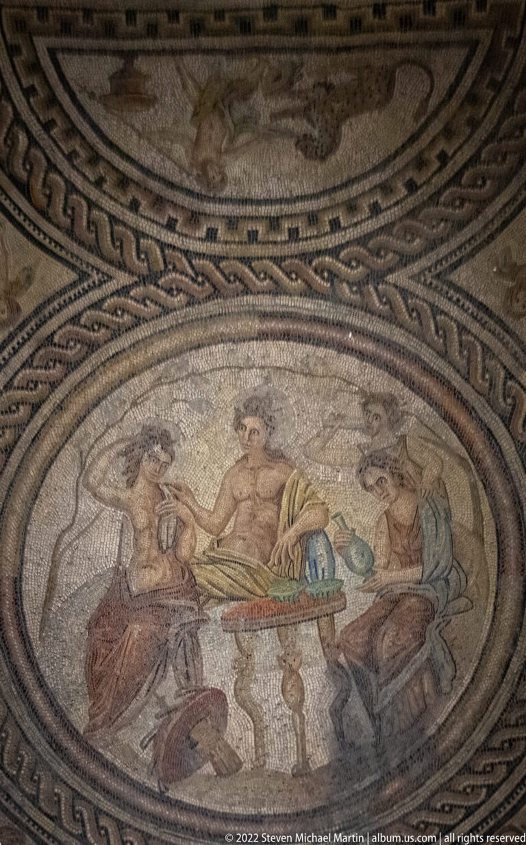 Circular Roman mosaic showing reclining figures around a central table, enclosed by braided and geometric borders.