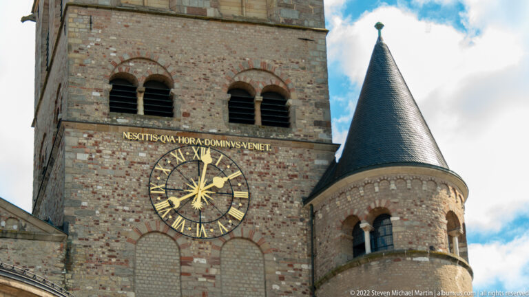 Gilded clock on Trier Cathedral masonry beside a round conical-roofed turret and Romanesque windows.