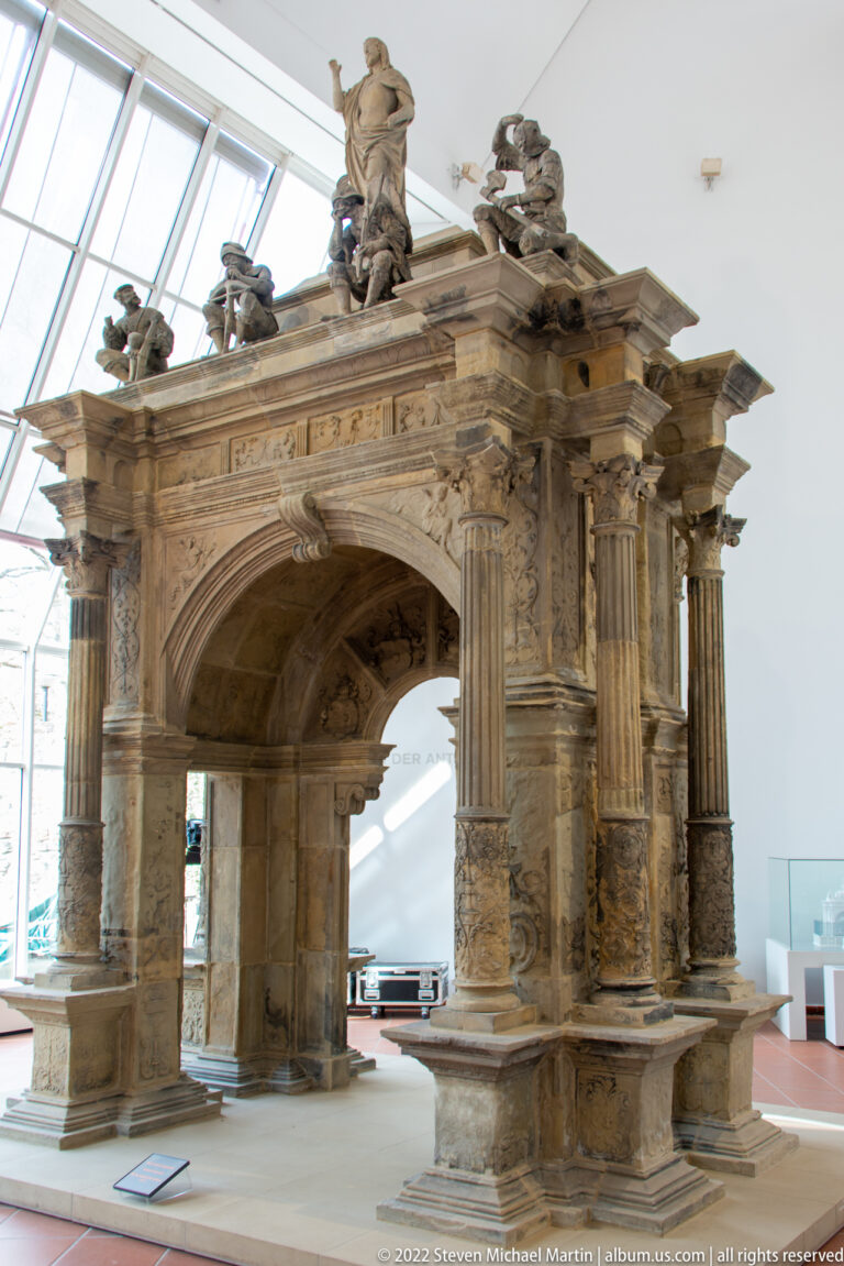 Freestanding reconstructed Roman stone monument with a central arch, engaged columns, relief panels, and figures on top.