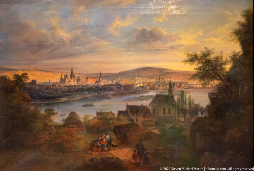 Painted panoramic view of Trier across a river valley, with church towers, foreground travelers, and a luminous cloudy sky.