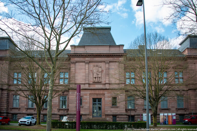 Symmetrical red-sandstone museum façade with central doorway, sculpted panel, bare trees, and a modern vertical sign.