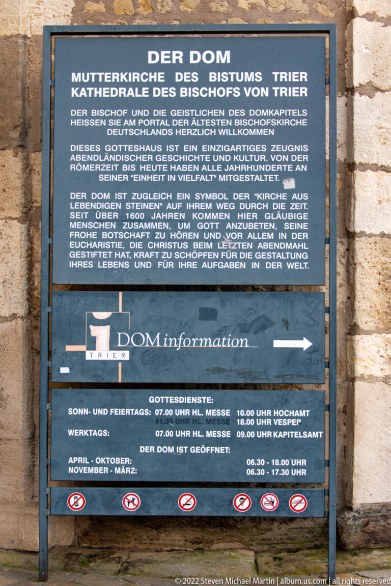 Large German information board for Trier Cathedral mounted on a stone wall, with text, service times, and directional graphics.