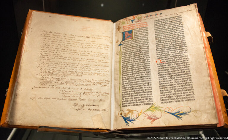 Open early printed book with narrow columns of black type, painted initial, colored pen flourishes, and handwritten annotations.