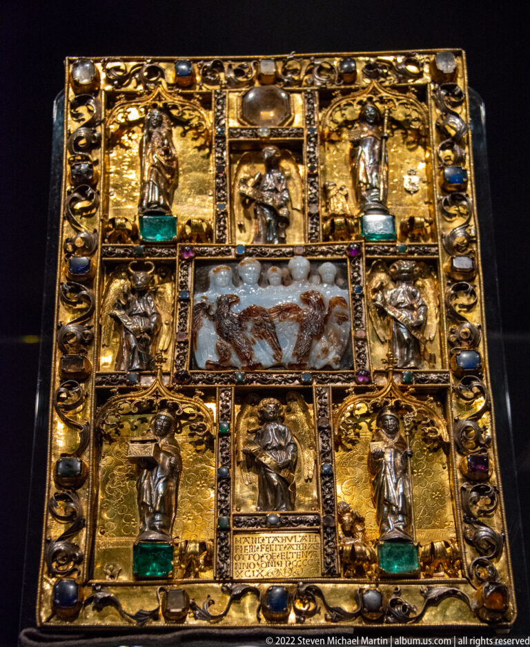 Square gilded medieval panel with small relief figures, gemstones, scrollwork, and a central translucent carved stone scene.