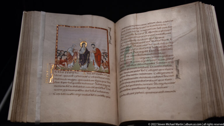 Open medieval manuscript with a small illuminated scene of haloed figures above Latin text on parchment.