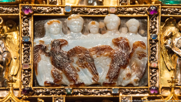 Translucent carved relief with human heads above large brown birds, set in a jeweled gilded frame.