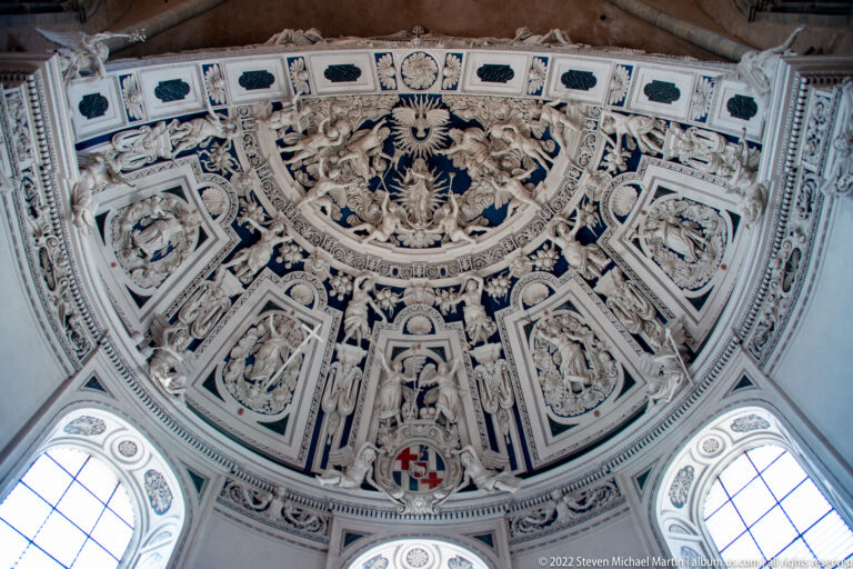 Elaborate white stucco ceiling with sculpted figures, heraldic emblems, framed reliefs, and windows below.