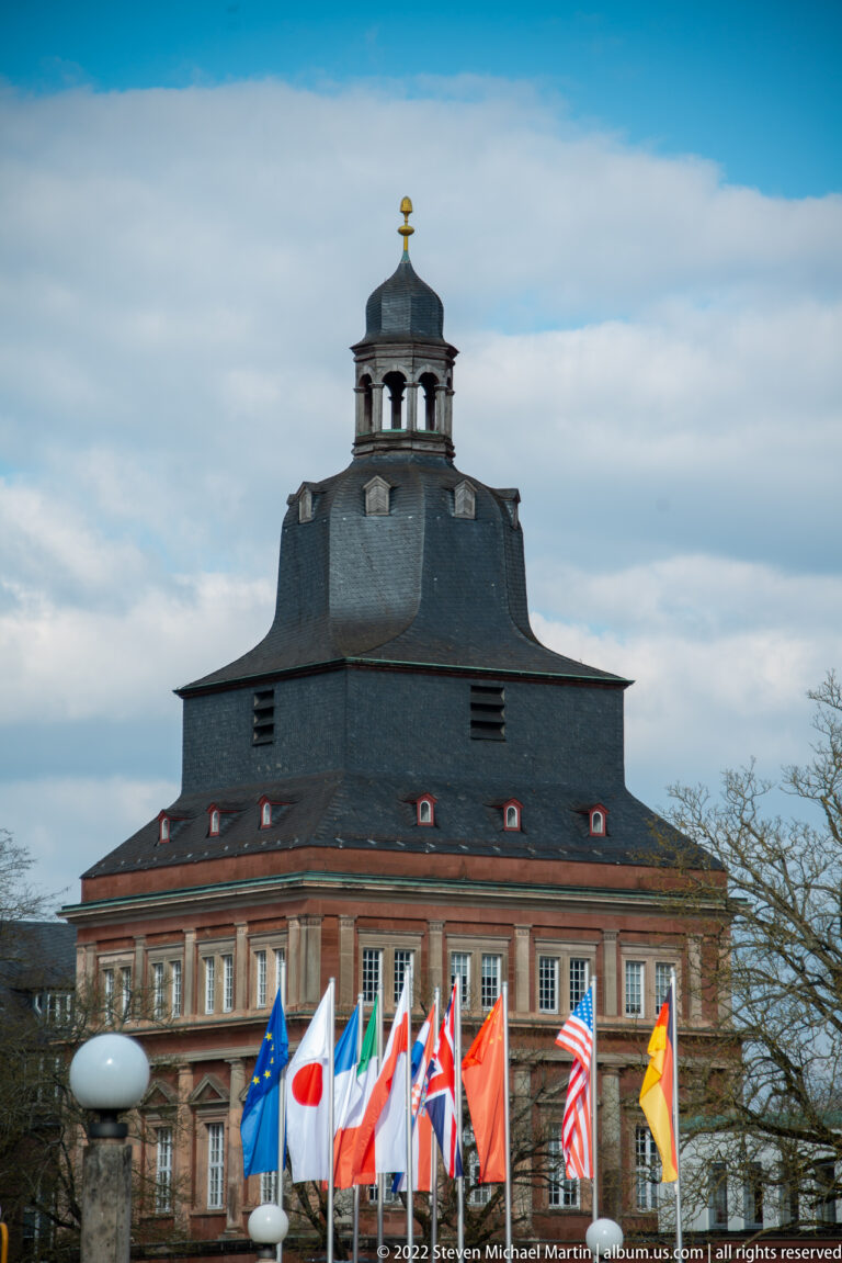 Historic red-and-cream building with a broad slate roof, octagonal cupola, and a row of international flags.