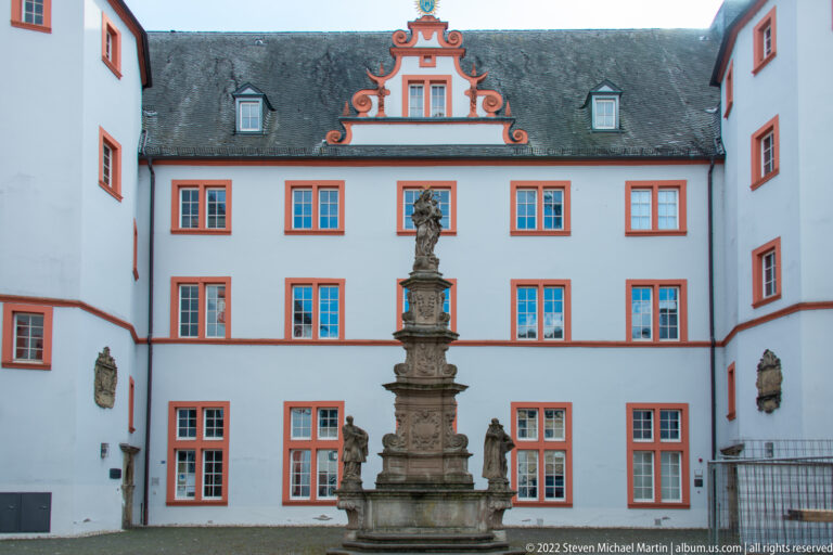 White-and-red historic courtyard with stepped gable and a tall carved stone fountain or monument at center.