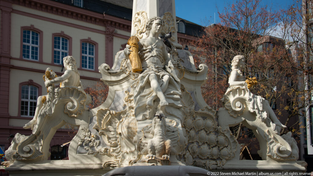 Ornate white stone Rococo fountain group with seated figures, curling scrolls, shells, and gilded accents.