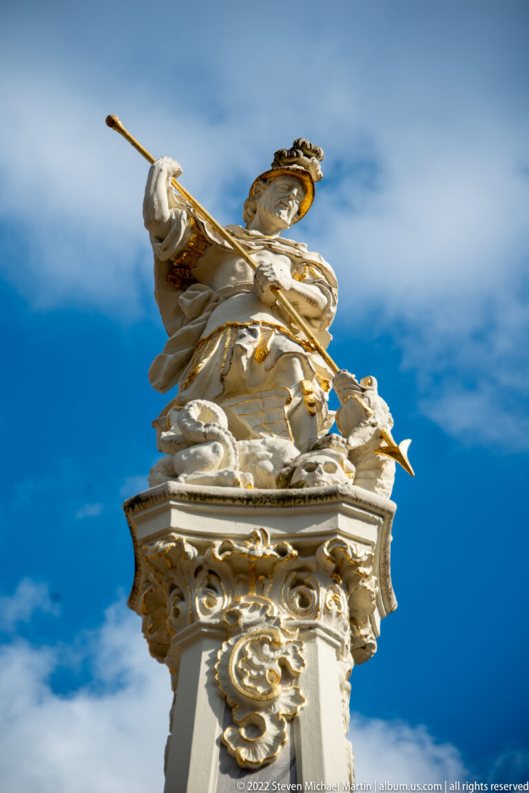 Low-angle view of the Georgsbrunnen's crowning figure with white stone drapery, gilded ornament, and blue sky.