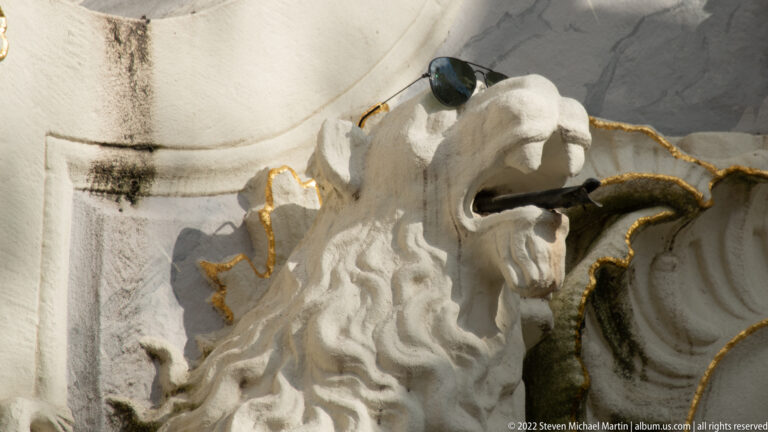 White carved lion head with dark metal water spout and gilded rocaille trim on an ornate fountain.