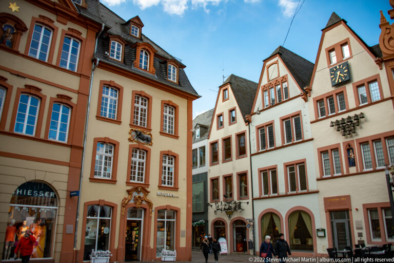 Colorful historic town houses with steep gables, a gable clock, shops, and pedestrians on Trier's market square.