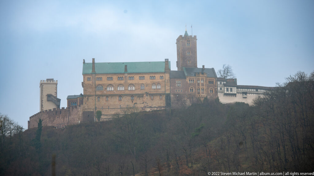 Wartburg Castle on a wooded hill, with long stone ranges and a tall central tower under a hazy sky.