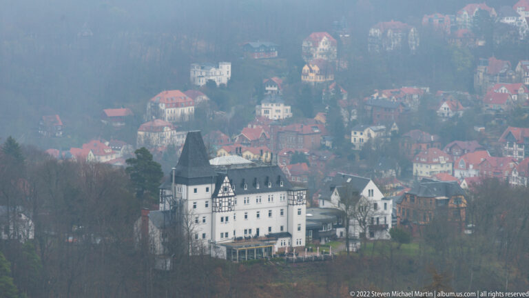 White multi-story historic building with dark roof on a wooded hillside below Wartburg, surrounded by houses in mist.