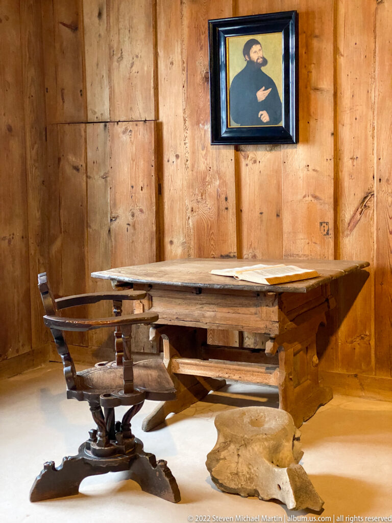 Wooden writing table and carved chair beneath a framed portrait of Martin Luther on paneled walls.
