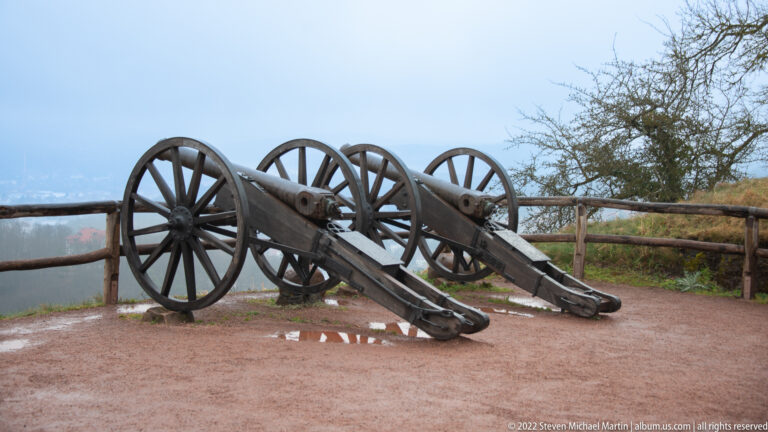 Two small wheeled cannon-like artillery pieces displayed on gravel beside a wooden railing and valley overlook.