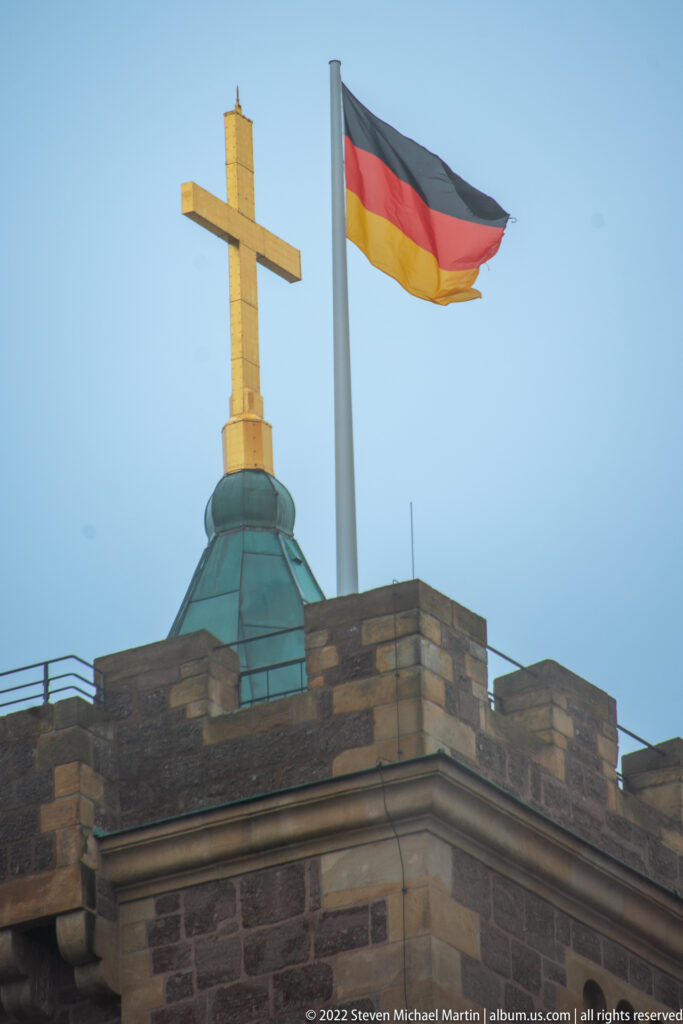 Large cross and German flag above a crenellated stone roofline at Wartburg.