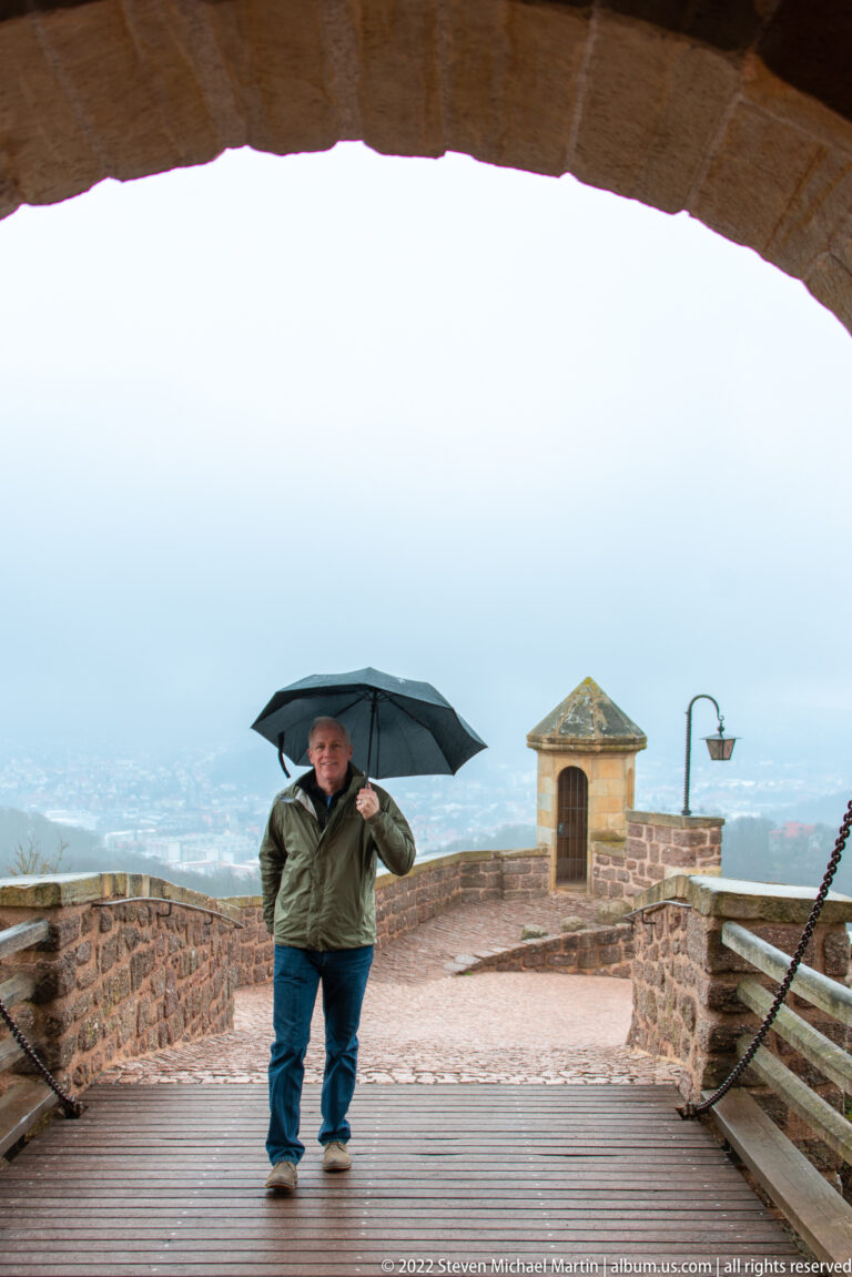 Visitor carrying a black umbrella on a wet Wartburg terrace, with parapet and hazy valley beyond.