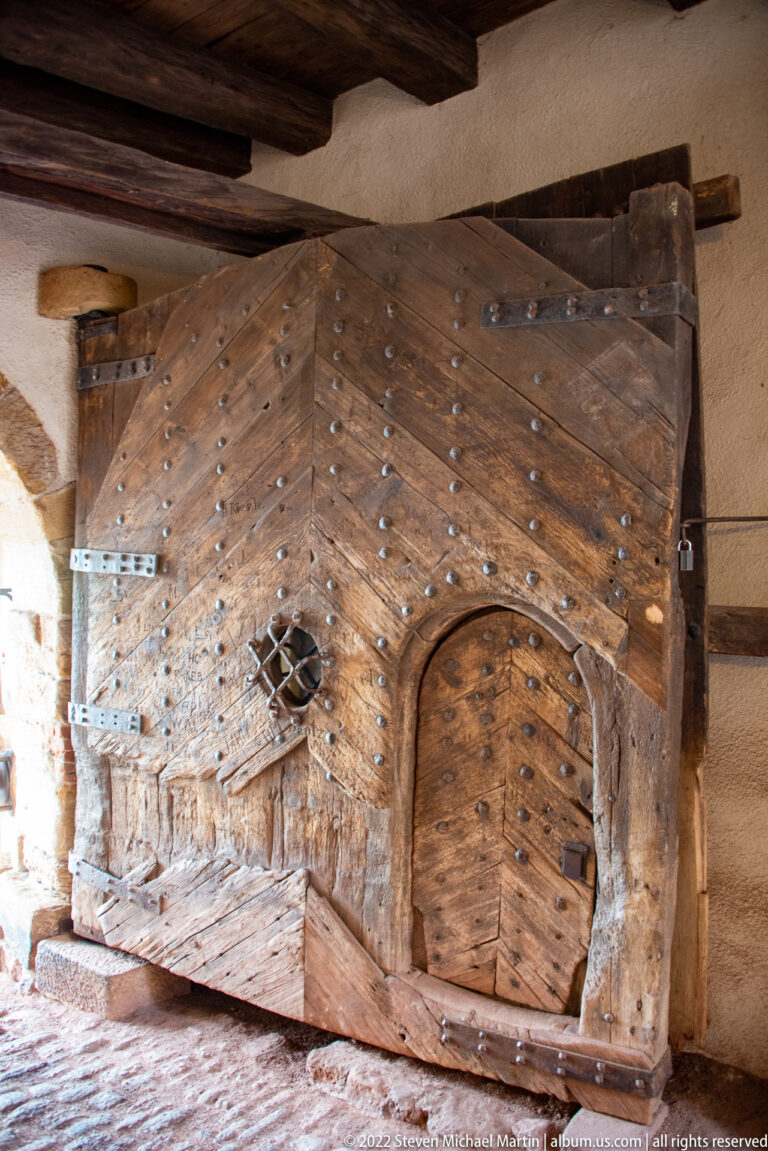 Large studded wooden gate with diagonal planks, heavy hinges, and a smaller rounded wicket door set into it.