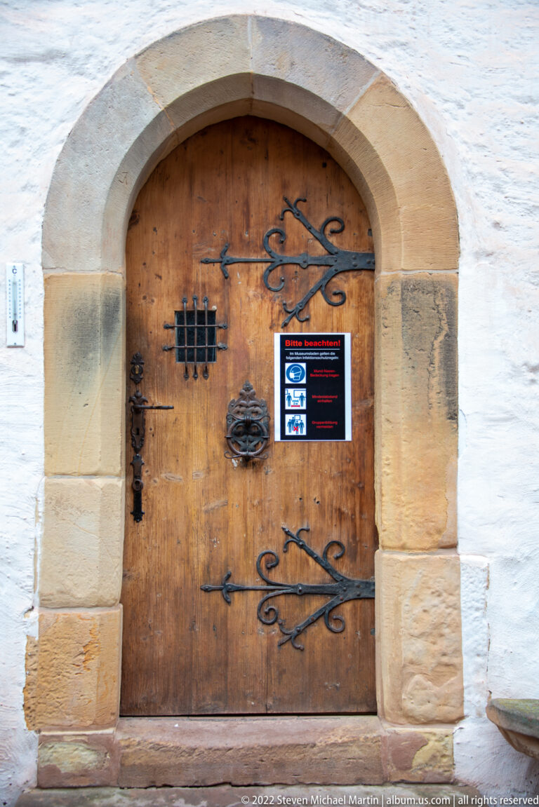 Arched wooden door in a stone surround with ornate black iron hinges, ring handle, barred opening, and small modern notice.