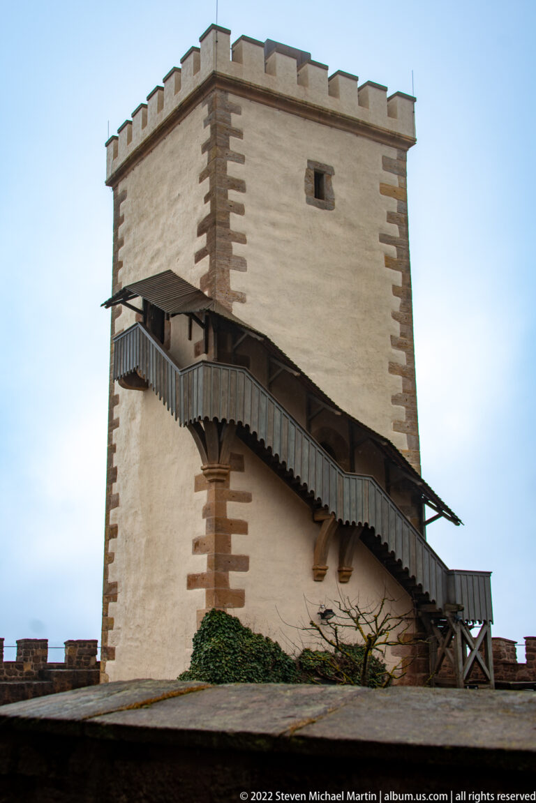 Square pale tower with stone corner blocks, crenellated top, and a covered exterior stair climbing along one side.