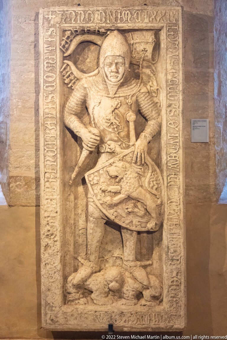 Carved stone slab with a standing armored figure, shield, border inscription, and small figure or animal at the feet.