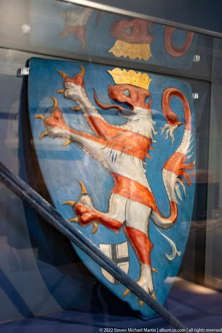 Red-and-white striped crowned lion rampant on a blue heraldic shield behind glass.