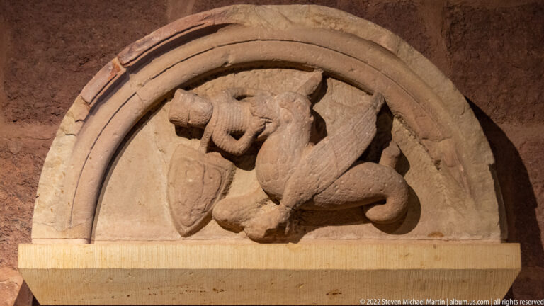Carved stone lion in relief beneath a semicircular arch, with curled tail and a shield-like form beside its body.