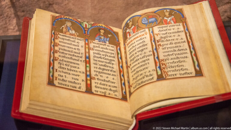 Open illuminated manuscript with medieval script, colored initials, and small painted figures displayed under glass.