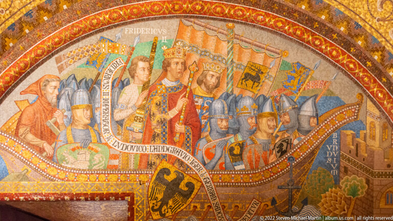 Crowned medieval ruler surrounded by attendants, soldiers, heraldic shields, and inscription bands in a Wartburg mural.