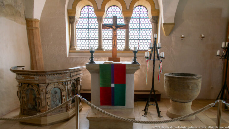 Small Wartburg chapel with altar and cross before arched windows, carved pulpit at left, and stone baptismal font at right.