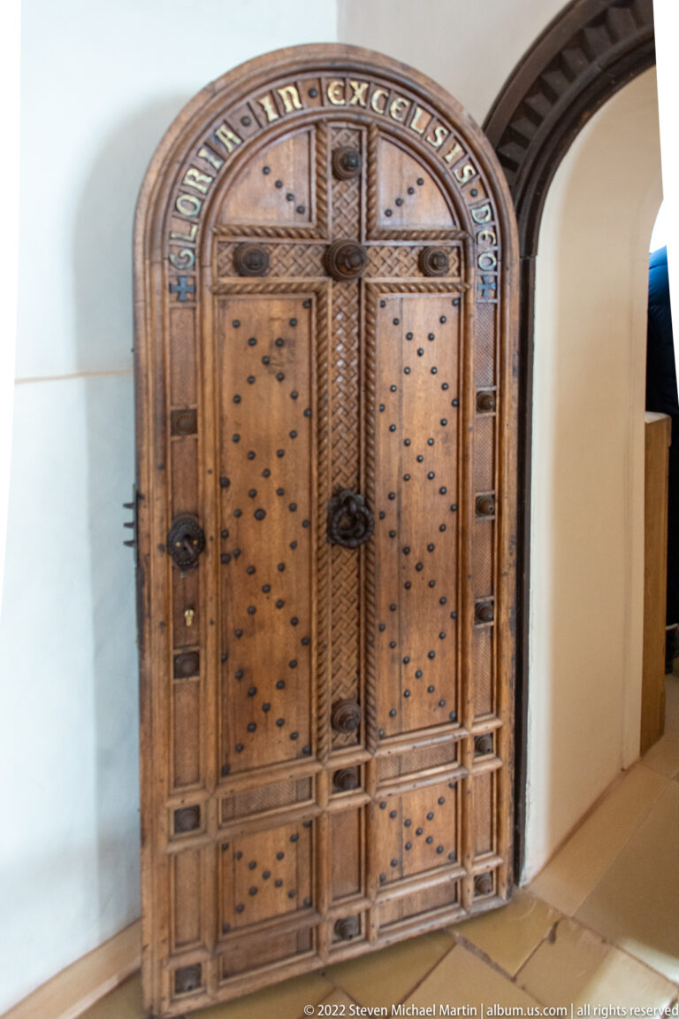 Tall arched wooden door with metal studs, carved geometric borders, heavy iron fittings, and lettering around the top.