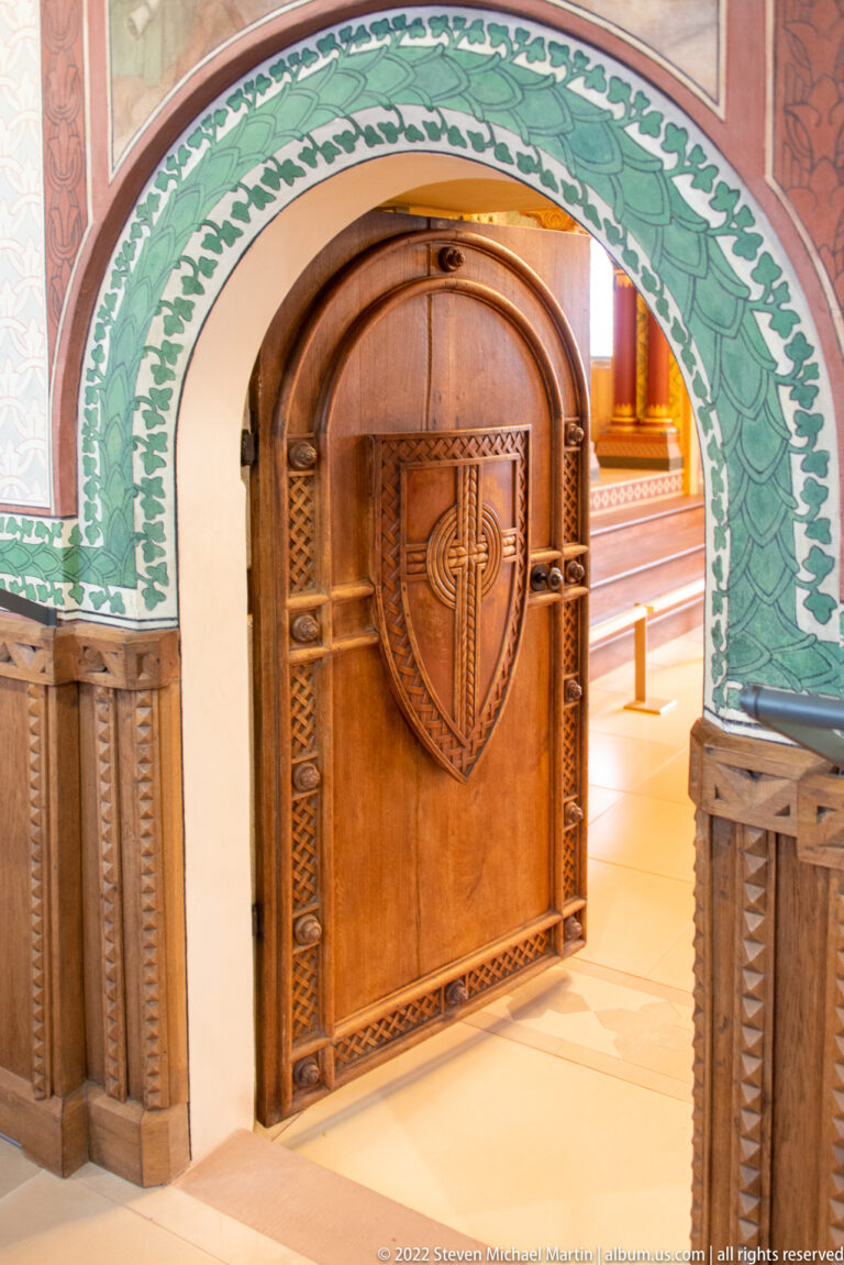 Rounded wooden door with carved heart-shaped panel beneath a green painted foliate arch and carved wall paneling.