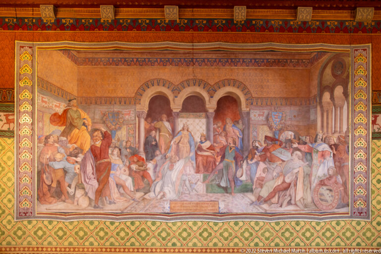 Large mural of a crowded medieval court scene inside an arcaded hall, surrounded by a patterned decorative border.