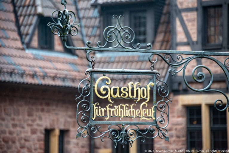 Decorative wrought-iron hanging sign reading 'Gasthof für fröhliche Leut' in front of a historic building.