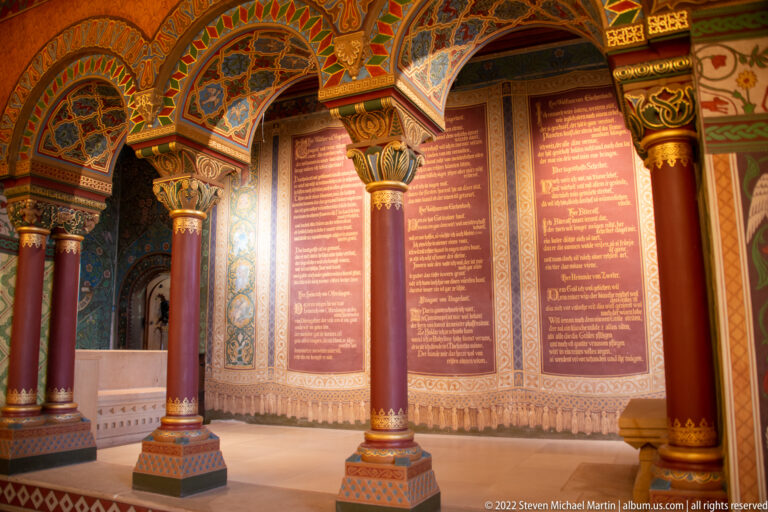 Decorated Wartburg hall with red columns, carved capitals, painted arches, and large inscribed wall panels.