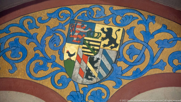 Quartered heraldic shield with lions, stripes, and diagonal bands surrounded by blue scrolling foliage on gold.