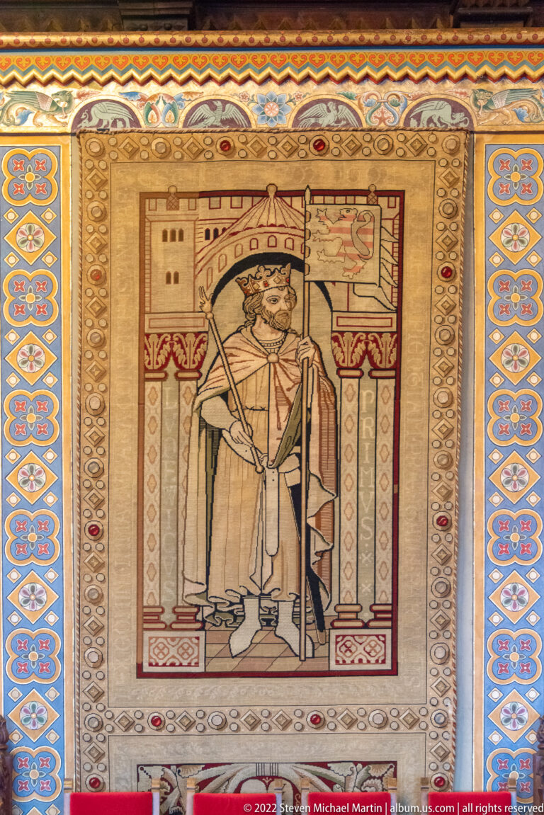 Standing crowned ruler holding a long ceremonial object beneath a canopy, surrounded by a patterned border.