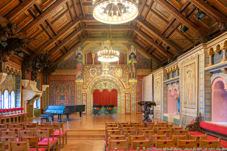 Long decorated hall with timber coffered ceiling, chandeliers, rows of chairs, murals, and ornate wall surfaces.
