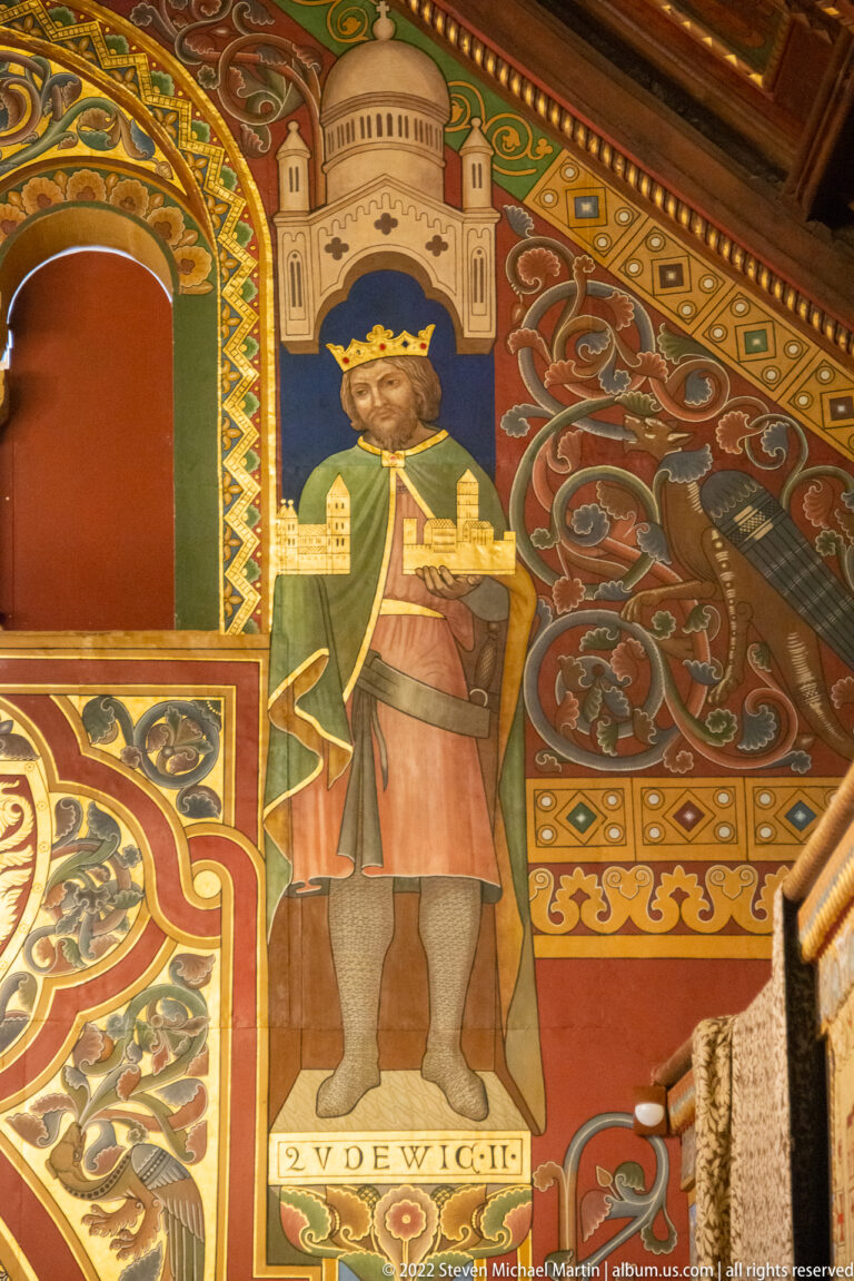 Painted crowned medieval ruler in green tunic holding a long sword against ornate red and gold decoration.