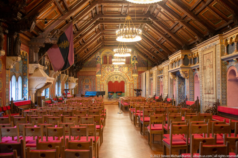 Axial view down a long Wartburg hall with repeated chandeliers, timber ceiling, ornate walls, and rows of red-seated chairs.